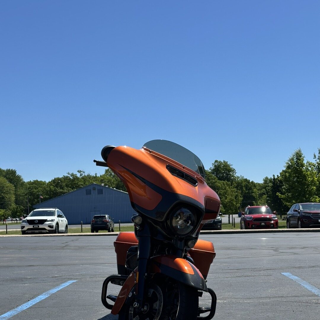 Orange sport motorcycle parked in a parking lot under a clear blue sky, front wheel turned, with a windshield and side panniers visible behind.