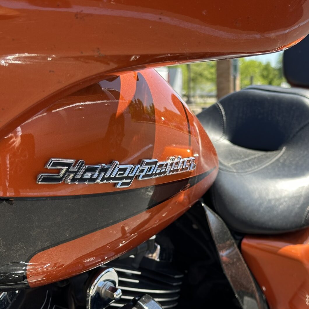 Close-up of an orange Harley-Davidson motorcycle fuel tank with a chrome emblem and black seat nearby.